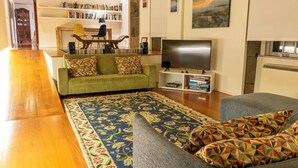 Una sala de estar con un sofá verde, un sofá gris, un televisor de pantalla plana, una estantería y piso de madera.