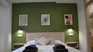 Cama bem arrumada com duas toalhas dobradas, cabeceira de madeira e três luminárias fixadas na parede.