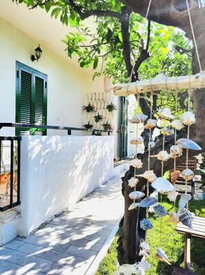 A white building with green shutters, a tiled patio, hanging plants, and a tree with hanging shells. - Il Canto delle Sirene VIESTE center, with garden, near Pizzomunno beach (Vieste)