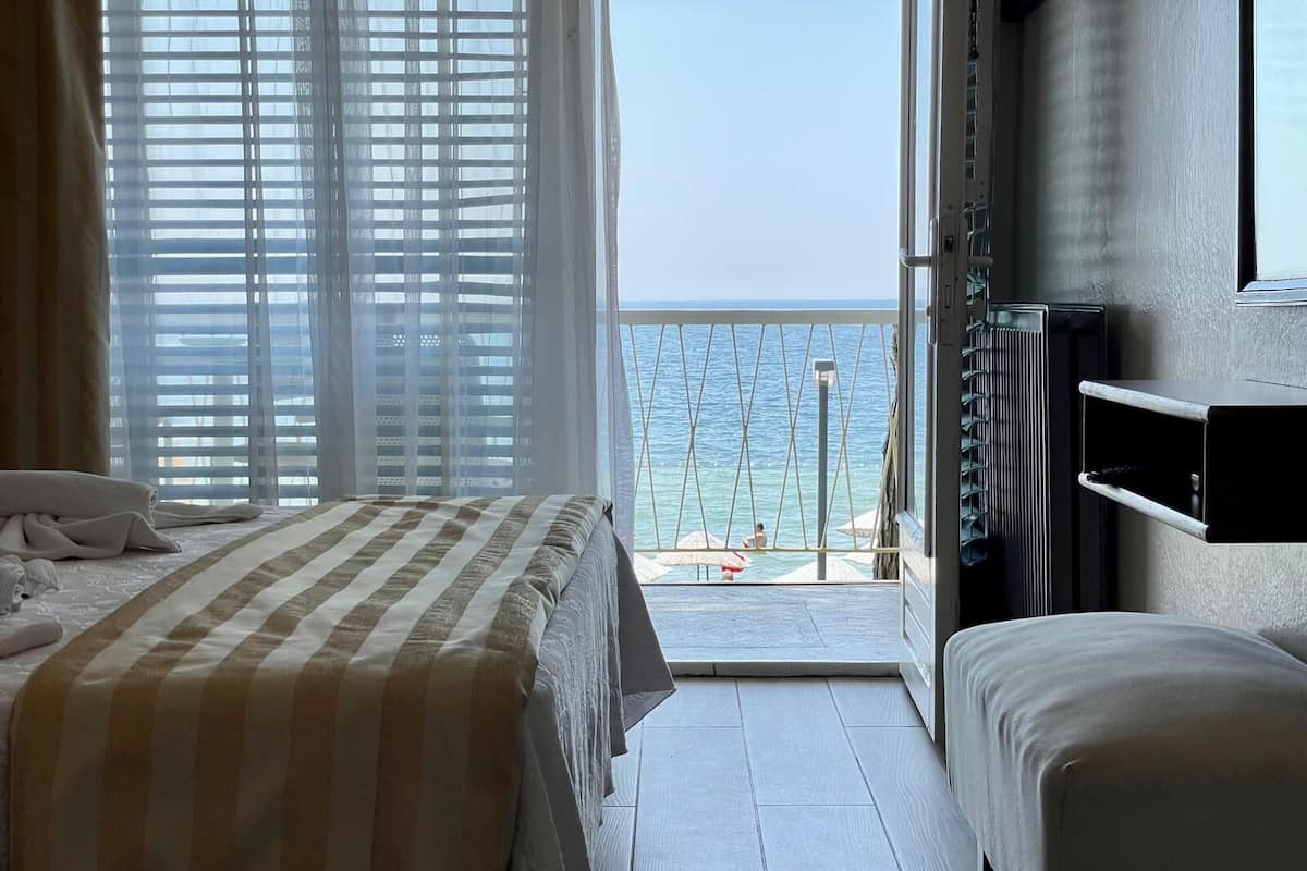 A hotel room with a bed, a small table, a TV, and a balcony with a view of the sea.