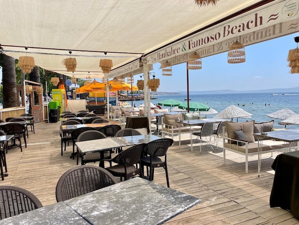 Ein Außenbereich zum Sitzen mit Tischen, Stühlen und Sonnenschirmen, mit Blick auf das Meer.