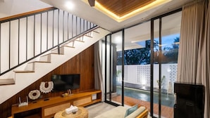 Ein modernes Wohnzimmer mit Treppe, Flachbildfernseher, Sofa und großem Fenster mit Blick auf einen Pool.