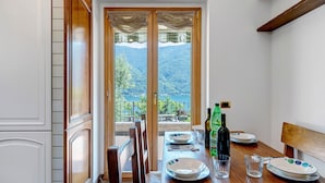 A dining area with a table set for two, a view of a mountainous landscape through a glass door, and wooden furniture. - Villa 'Porlezza Lake View' with Lake View, Air Conditioning, Private Pool, Ultra-Fast Wi-Fi (Cima)