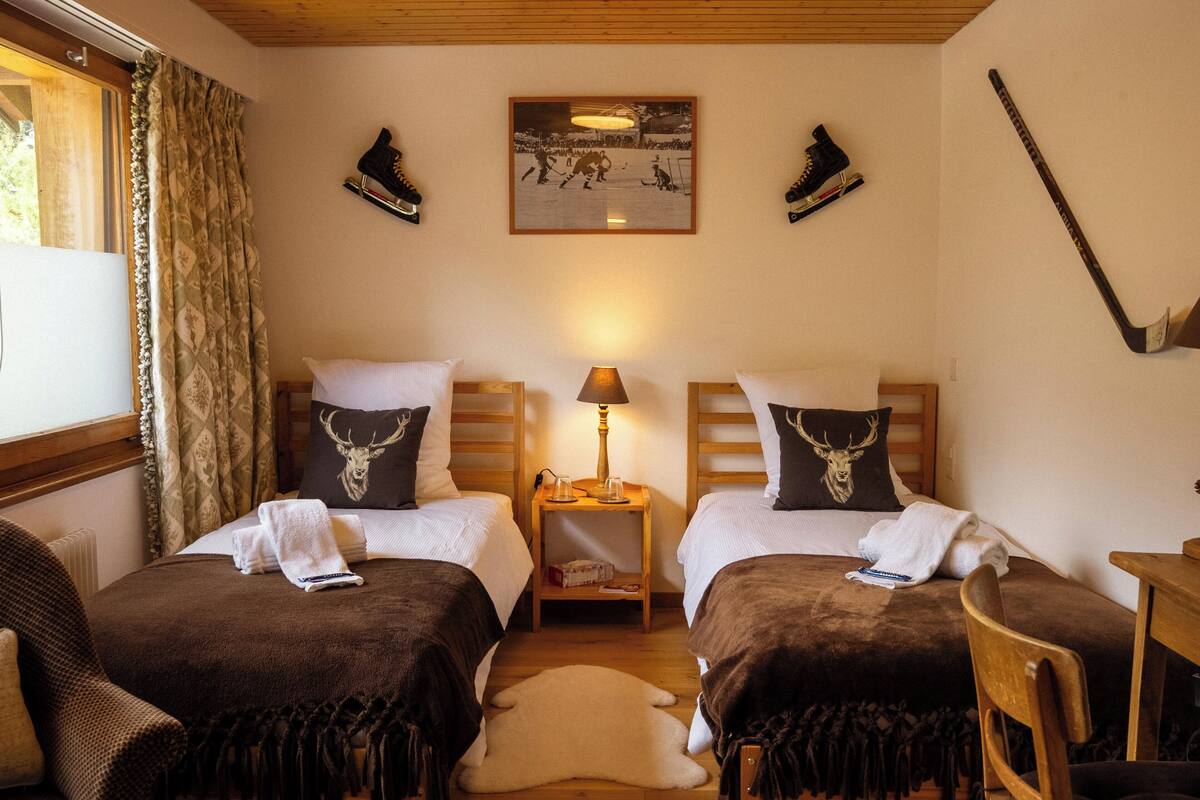 Ein Zimmer mit zwei Betten, einem Holzkopfteil, einer Lampe, einem gerahmten Bild an der Wand und einem an die Wand gelehnten Eishockeystock.