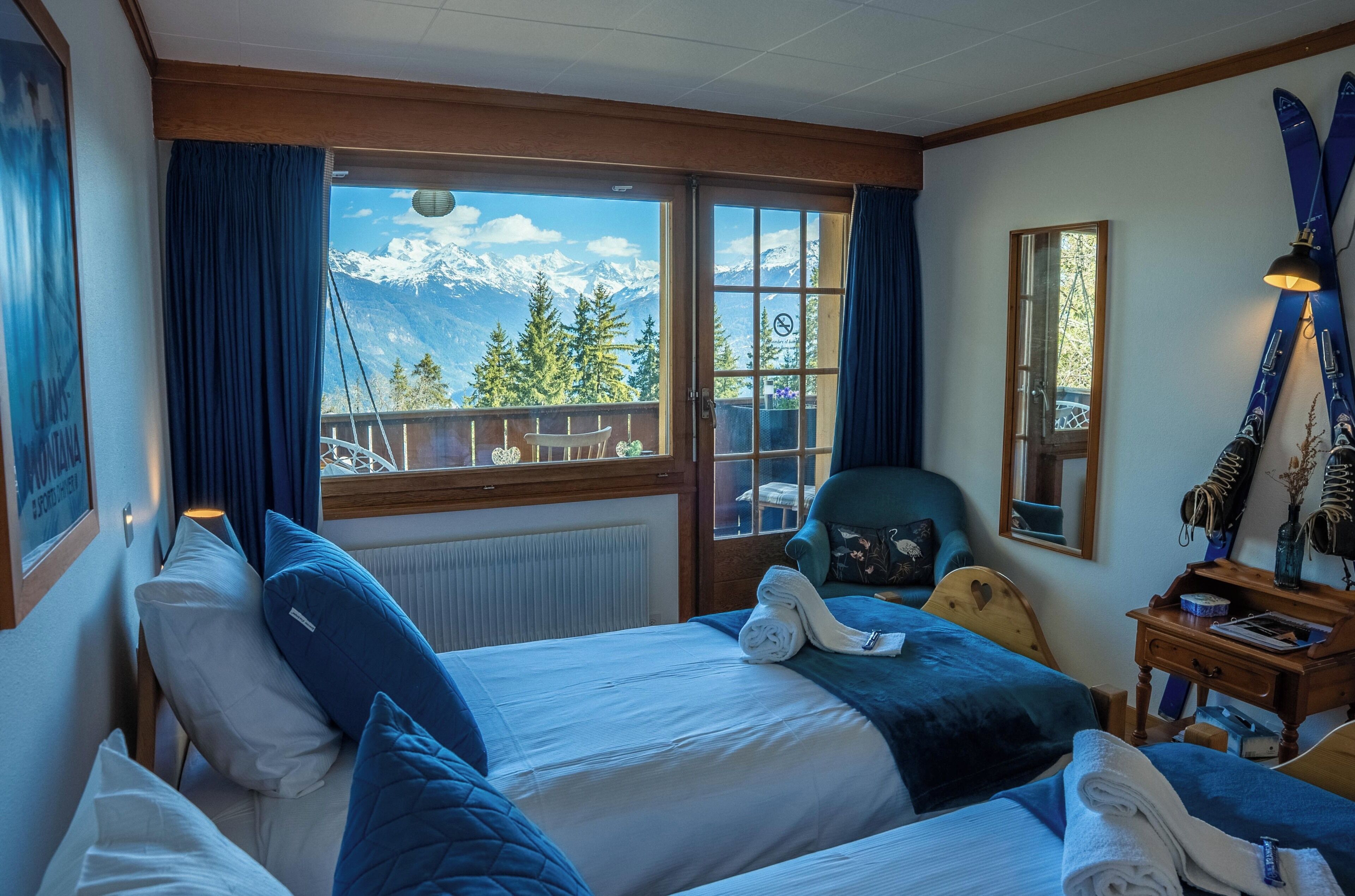 Camera d'albergo con due letti, vista sulle montagne innevate, soffitto in legno e finestra con tende.