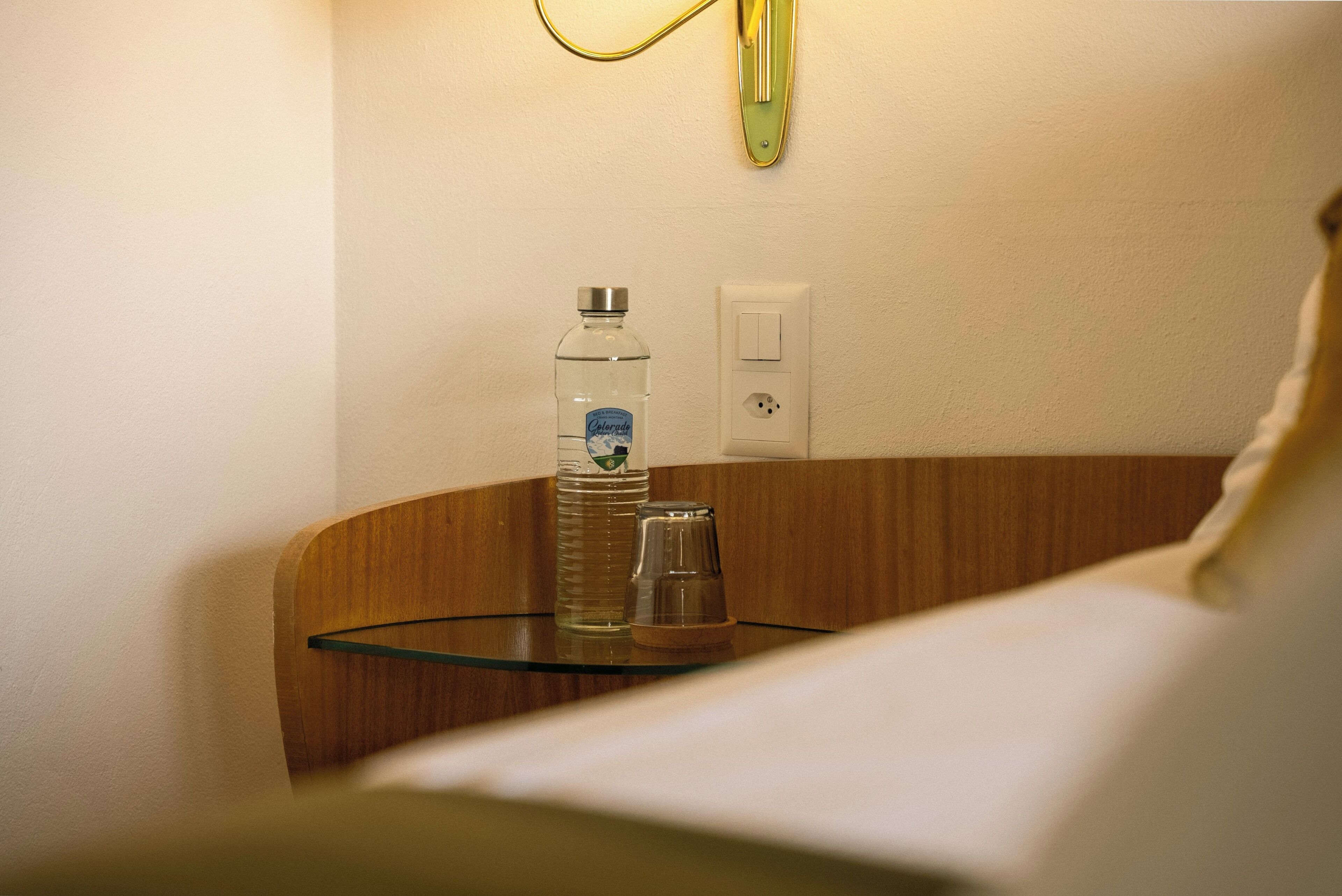 A hotel room with a glass table, a bottle of water, and a light switch on the wall.