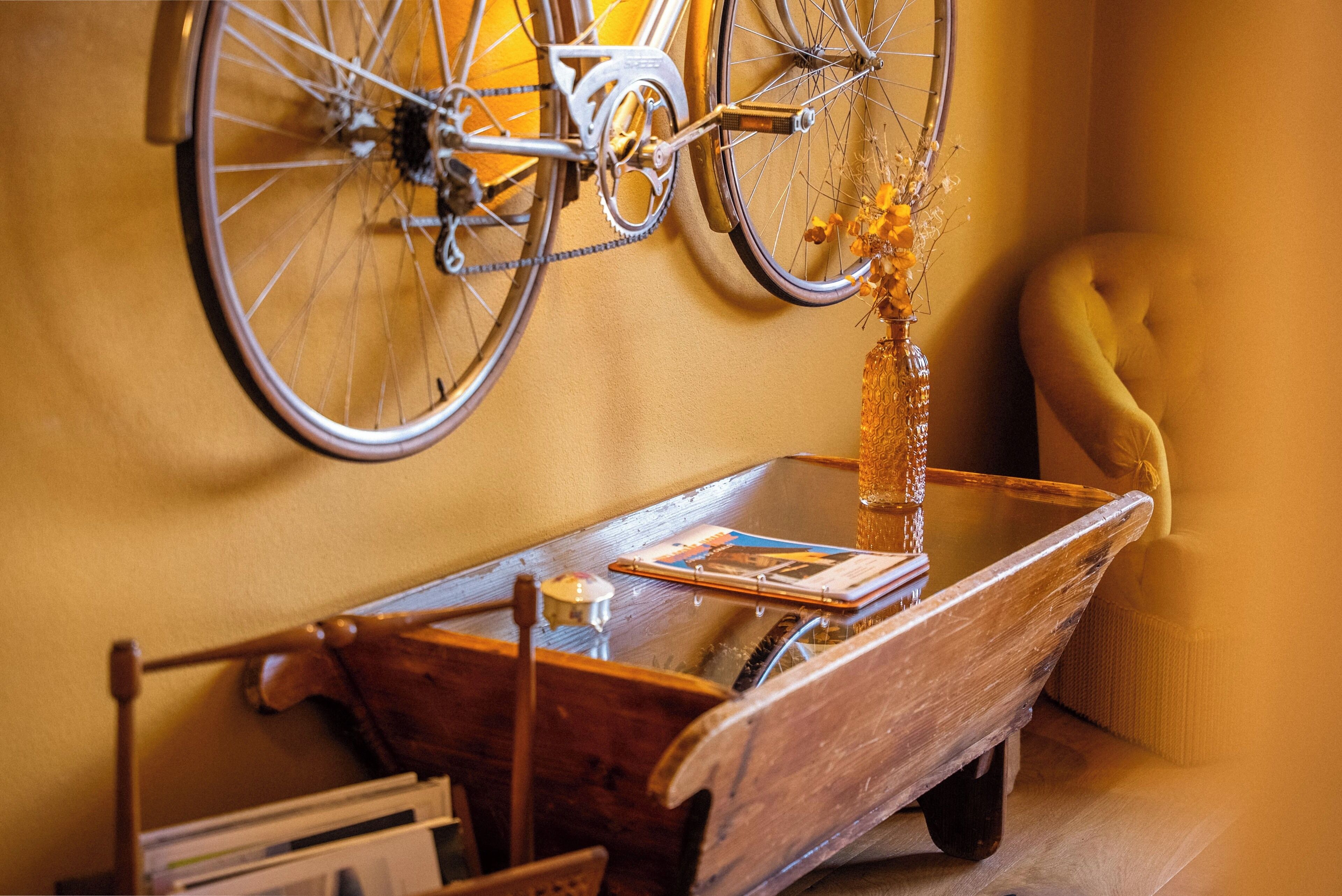 Een fiets aan een muur, een houten kar met een bloevas, en een gele fauteuil.