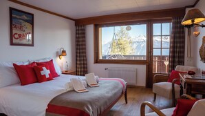 A hotel room with a bed, a table, chairs, a window with a view of mountains, and a ski lift poster on the wall.