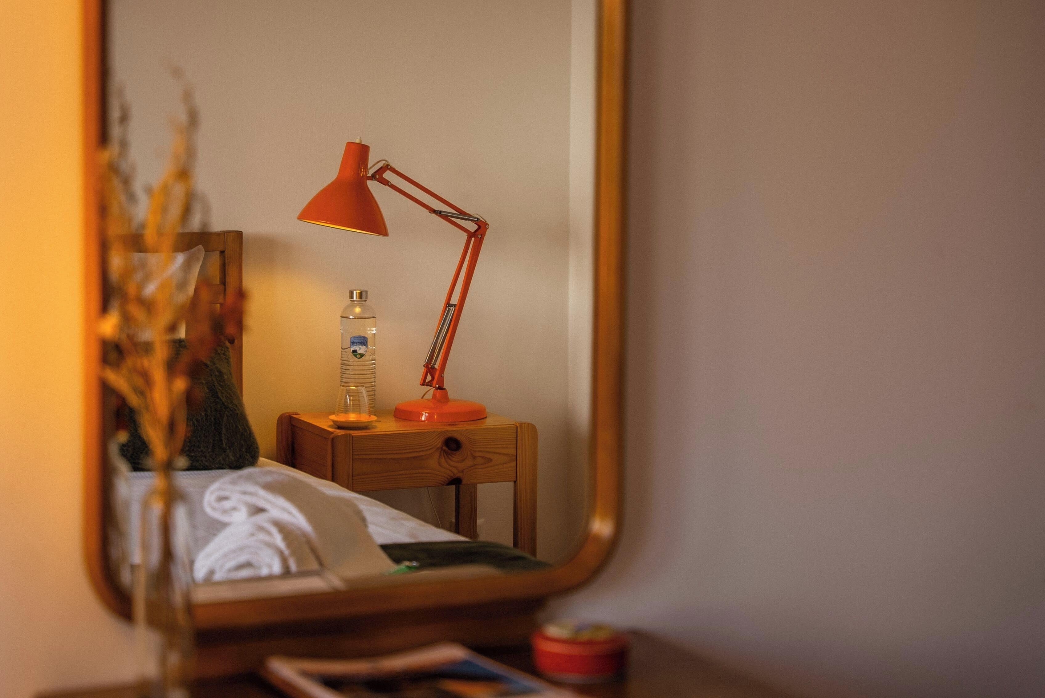 A mirror reflecting a bedside table with a lamp, a bottle, and a tissue box.