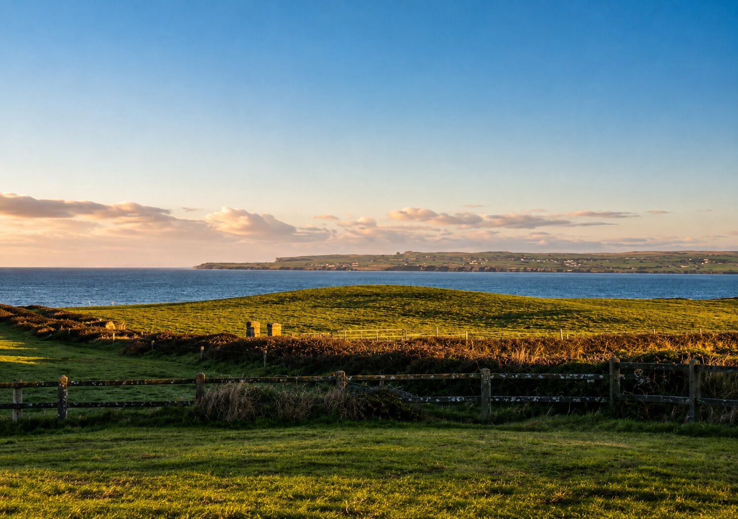 A coastal landscape with a grassy hill, a wooden fence, and a view of the sea and distant land.