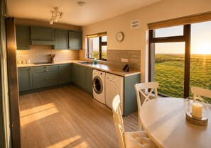 A kitchen with a washing machine, a dining table with chairs, a window with a view of the countryside, and a clock on the wall.