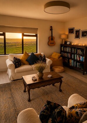 Una sala de estar acogedora con sofá, mesa de centro y estantería, una guitarra en la pared, y vistas a un campo a través de amplias ventanas.