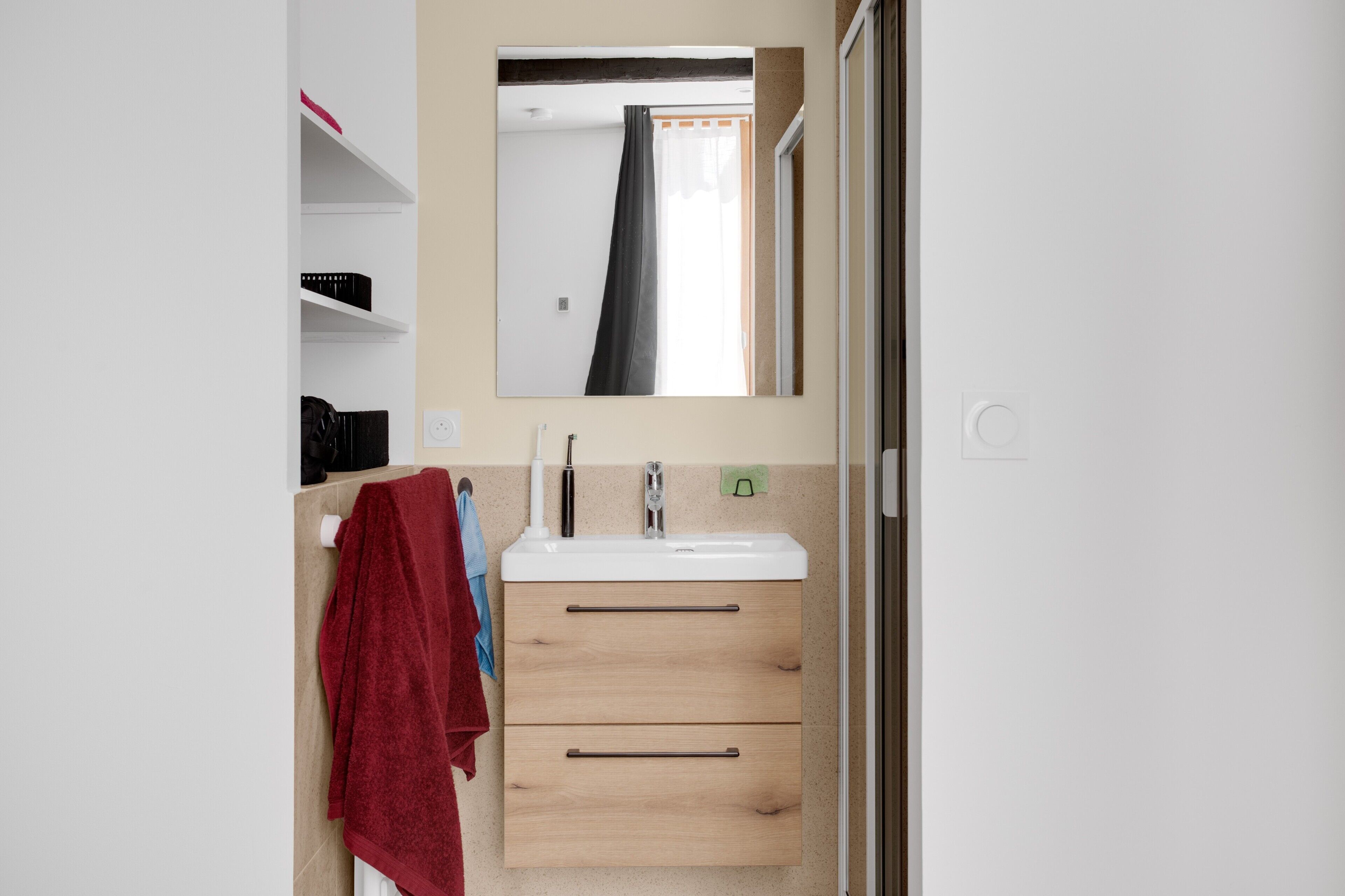 A bathroom with a white sink, wooden cabinet drawers, a towel rack with a red towel, and a mirror reflecting a window with a curtain.