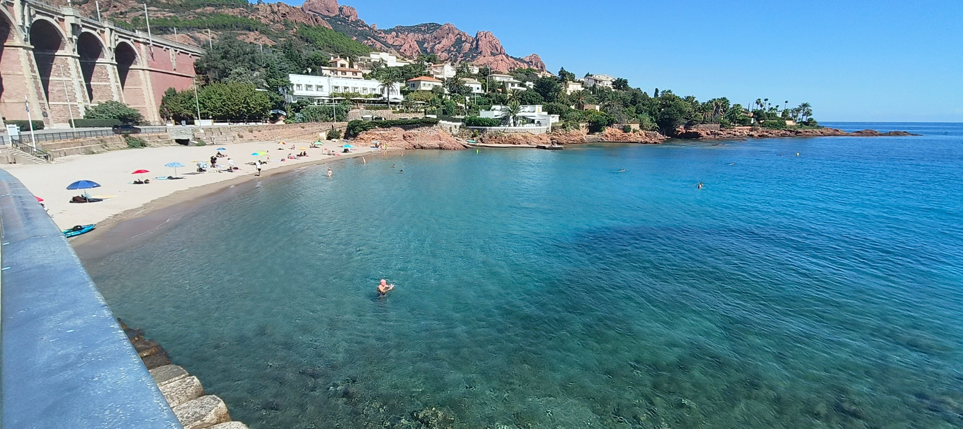 A coastal beach with clear water, a rocky shoreline, and buildings in the background.