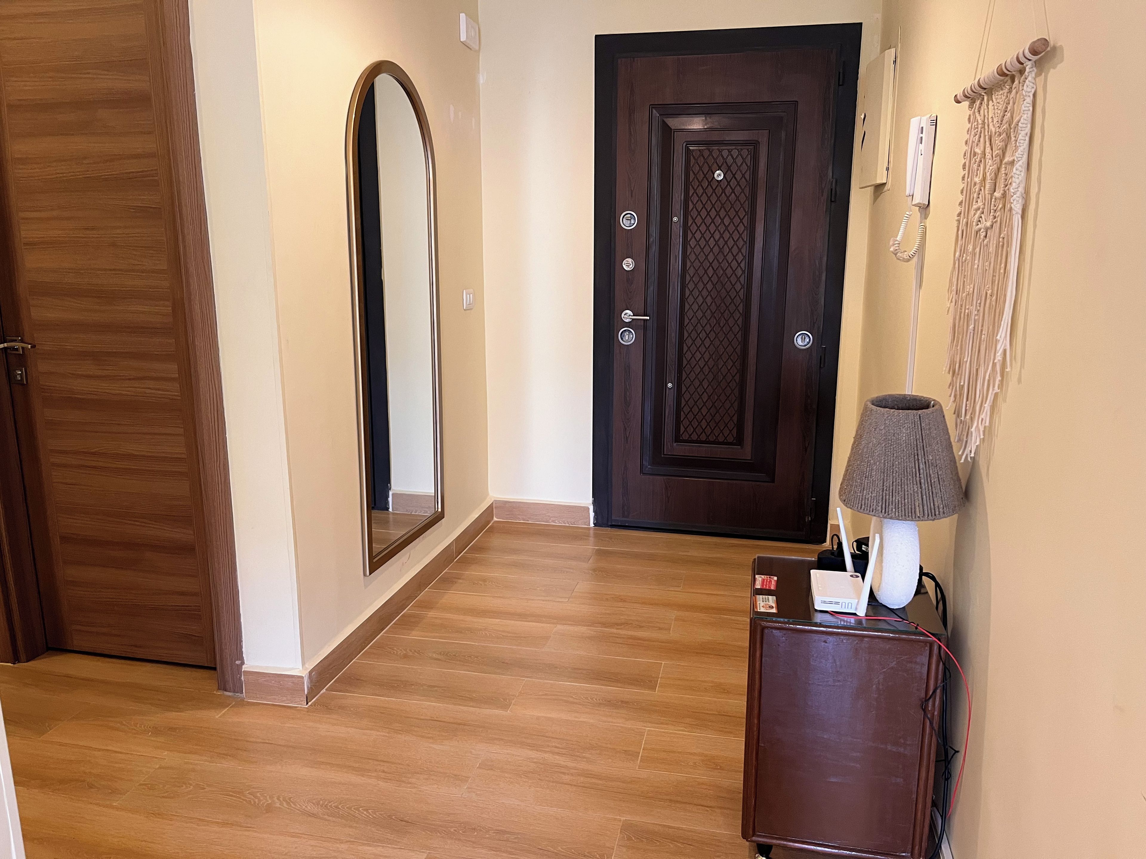 Un pasillo con suelo de madera, una puerta de madera oscura, un espejo grande y una lámpara sobre un mueble.