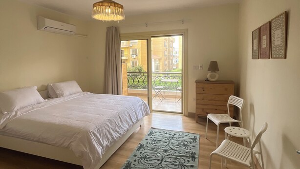 A bedroom with a bed, a dresser, a chair, a rug, and a view of the outside. - Spacious 3-bedroom apartment in the heart of Sheikh Zayed with Garden view (Giza Governorate)