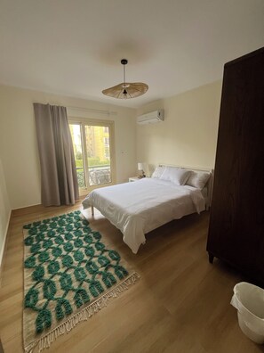 A bedroom with a bed, a green patterned rug, a wooden floor, a window with curtains, and an air conditioning unit. - Spacious 3-bedroom apartment in the heart of Sheikh Zayed with Garden view (Giza Governorate)
