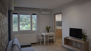 Una sala moderna con un sofá, una mesa de comedor con sillas, un televisor sobre un mueble bajo y un ventanal con vistas a la vegetación.