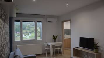 Una sala moderna con un sofá, una mesa de comedor con sillas, un televisor sobre un mueble bajo y un ventanal con vistas a la vegetación.
