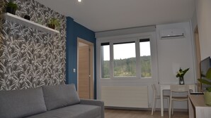 Una habitación con un sofá gris, un escritorio blanco con dos sillas, una ventana con vista a la vegetación y una pared con papel tapiz estampado con hojas.