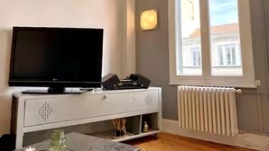 Quarto com televisão sobre um armário branco, uma janela com persianas e um radiador.