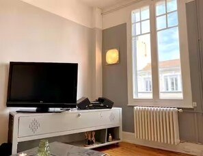 Une pièce avec un téléviseur posé sur un meuble blanc, une fenêtre avec des stores et un radiateur.