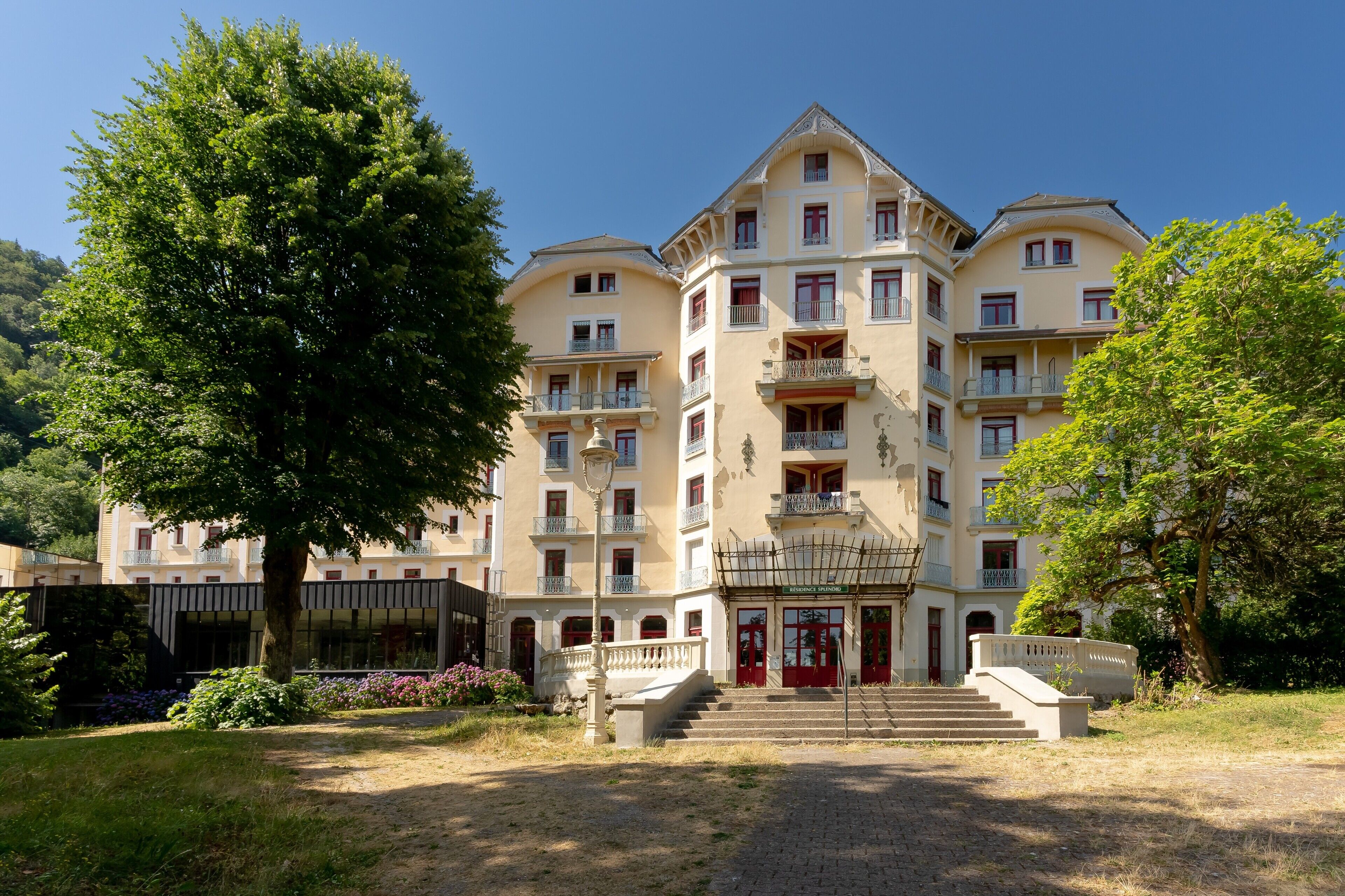 Un hotel a più piani con un ingresso centrale, balconi e un giardino circostante.