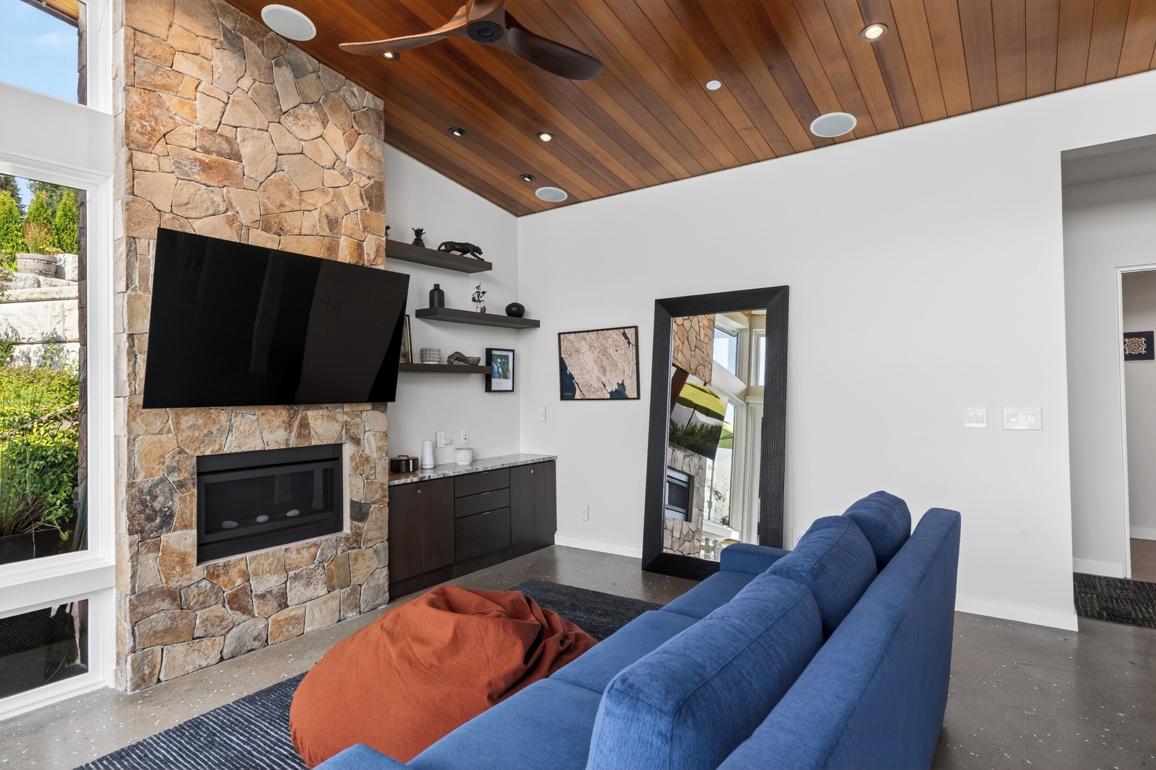 A modern living room with a stone fireplace, a flat-screen TV, a blue sofa, and a wooden ceiling.