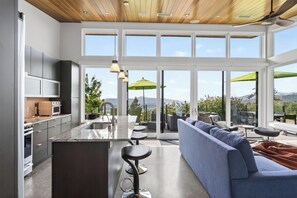 Eine moderne Küche mit einer zentralen Kochinsel, einem Essbereich mit einer Couch und Sesseln sowie großen Fenstern mit Blick ins Freie.