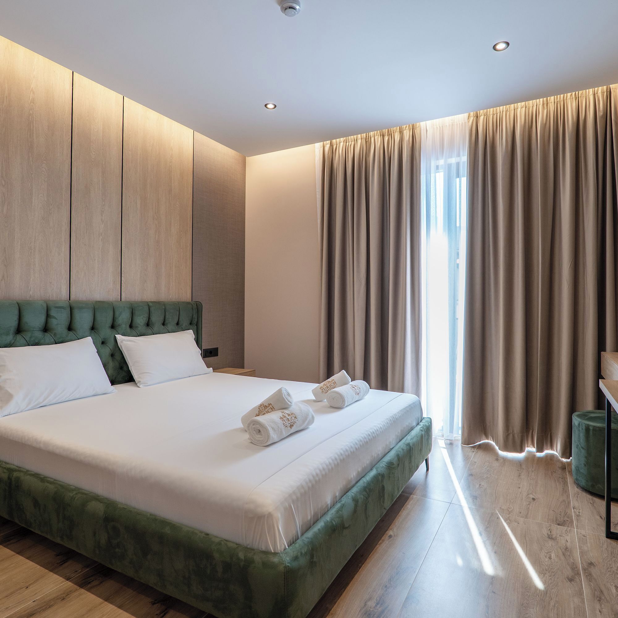 Une chambre d’hôtel moderne avec un grand lit, une tête de lit recouverte d’un tissu vert et une vue sur l’extérieur.