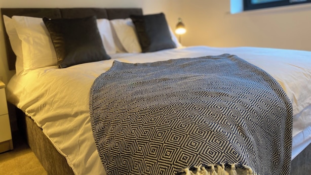 Sängyssä on kuviollinen päiväpeite, pääty ja seinään kiinnitetty lamppu.