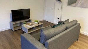 Olohuoneessa on taulutelevisio, sohvapöydällä on kulho täynnä hedelmiä, huoneessa on harmaa sohva ja seinällä taulu.