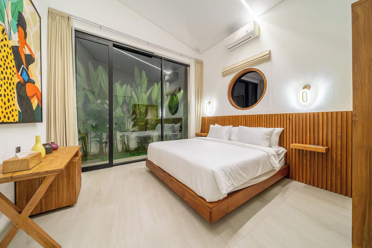 Un dormitorio moderno con cama, cabecera de madera, mesita de noche, espejo y puerta corrediza de vidrio que da a un balcón con plantas.