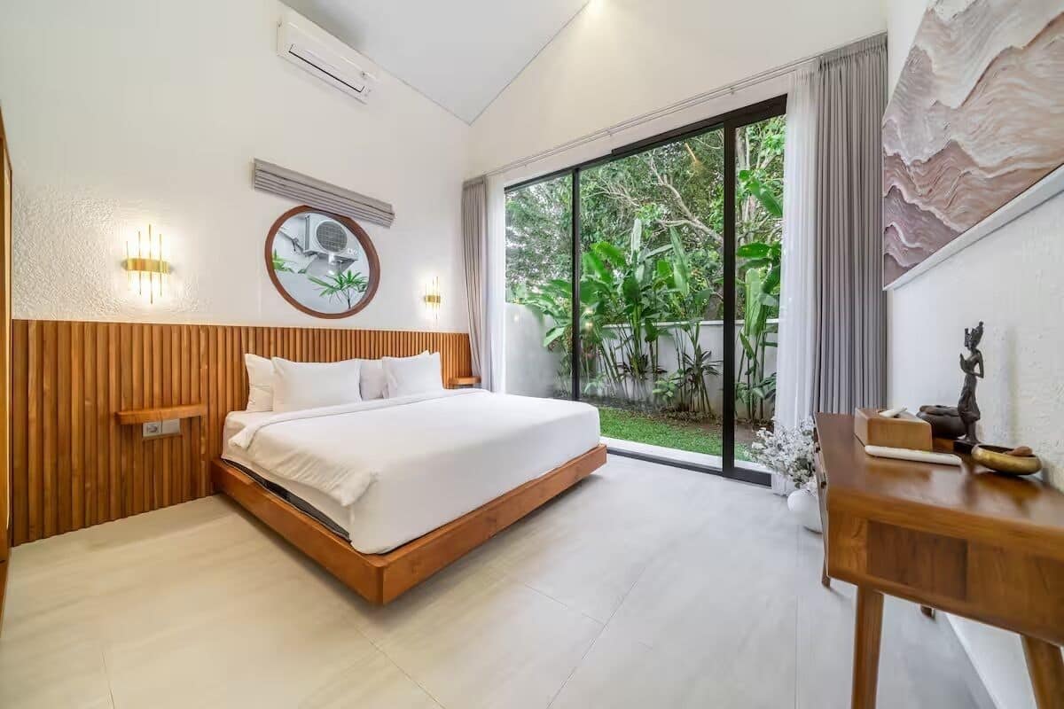 Un dormitorio moderno con una cama grande, paneles de madera, un espejo redondo, luces empotradas en la pared y vistas a la vegetación a través de una puerta corredera.