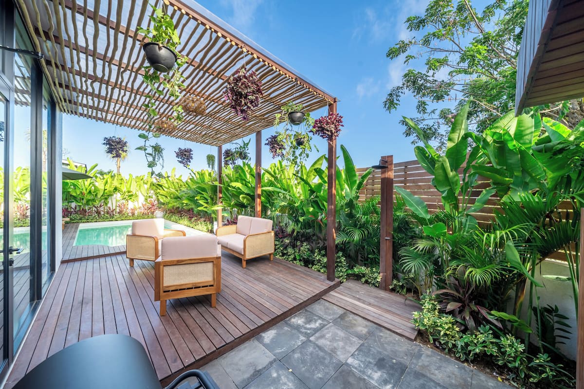Una terraza de madera con mobiliario exterior, una pérgola con plantas colgantes, una piscina y exuberante vegetación tropical.