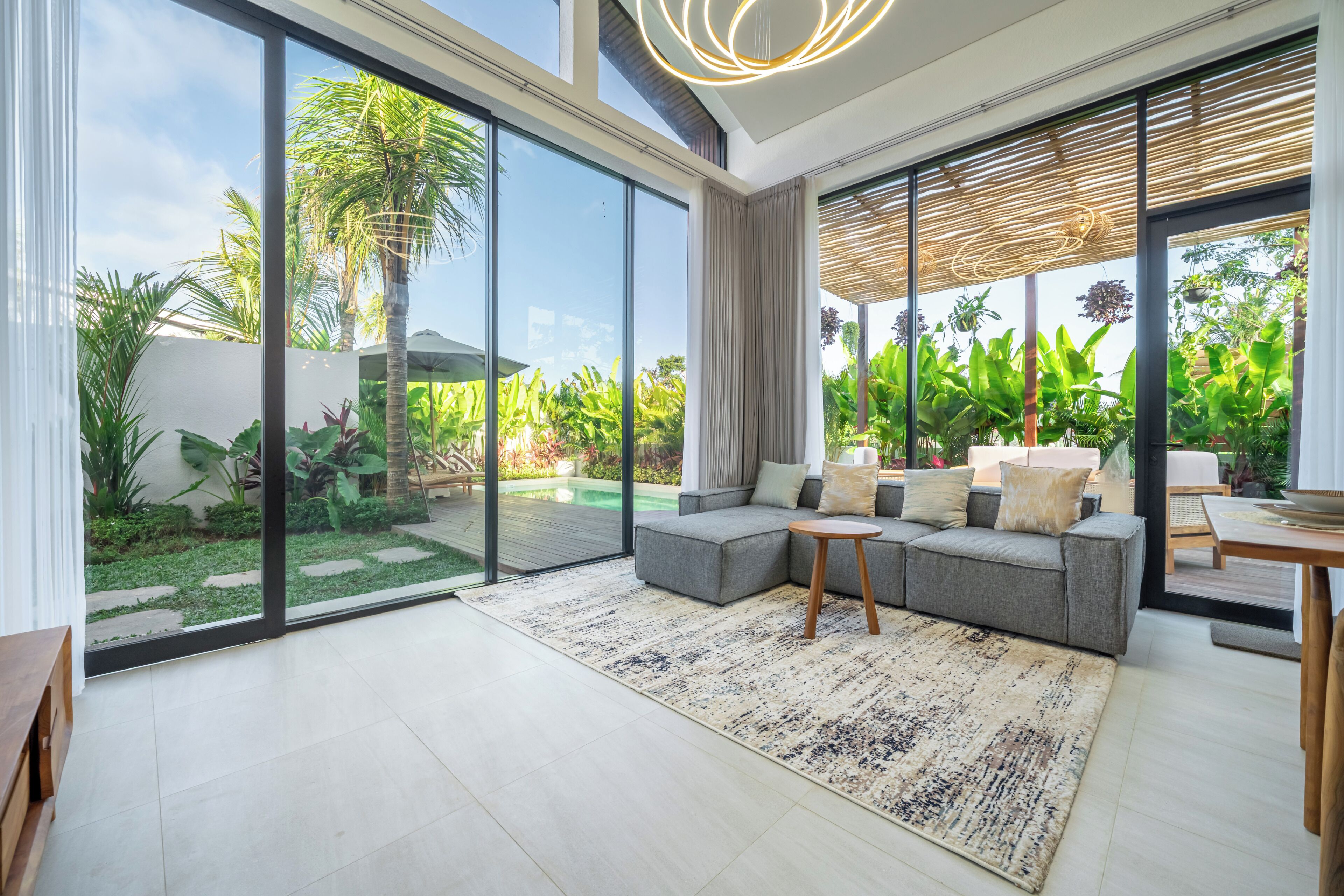 A modern living room with a grey sectional sofa, a small wooden side table, and a large glass sliding door leading to a garden with tropical plants.