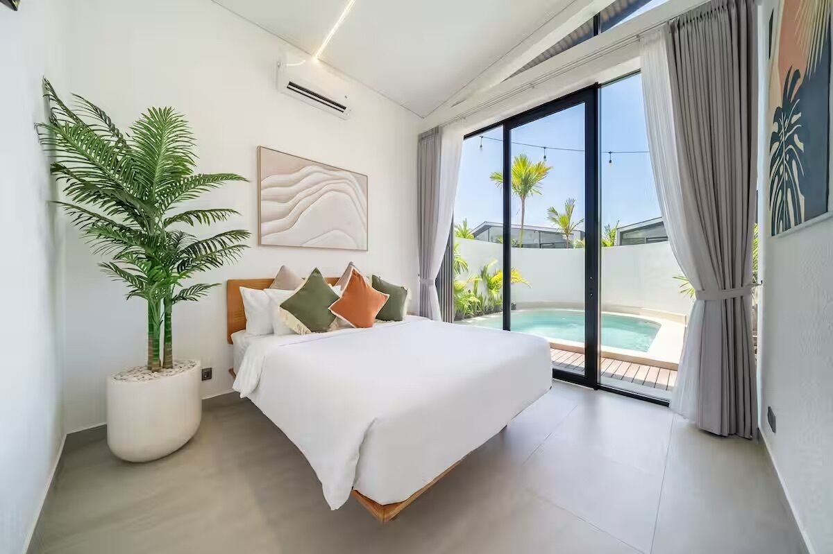Un dormitorio moderno con cama, una planta grande en maceta, un cuadro y vistas a una piscina.