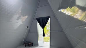 Quarto em formato triangular com uma cama, uma mesinha e uma janela com cortinas.