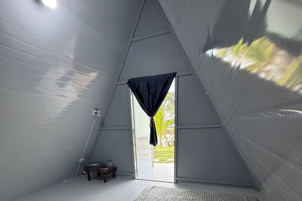 Una habitación triangular con una cama, una mesita pequeña y un ventanal con cortinas.
