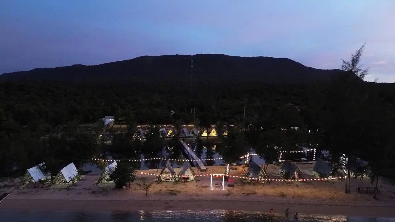 A beachside resort with multiple thatched-roof huts, a swimming area, and a view of a mountain range.