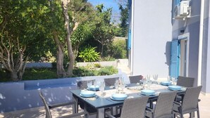 Ein Essbereich im Freien mit einem für sechs Personen gedeckten Tisch, umgeben von Korbstühlen, vor einer weißen Gebäudefassade mit blauen Fensterläden.
