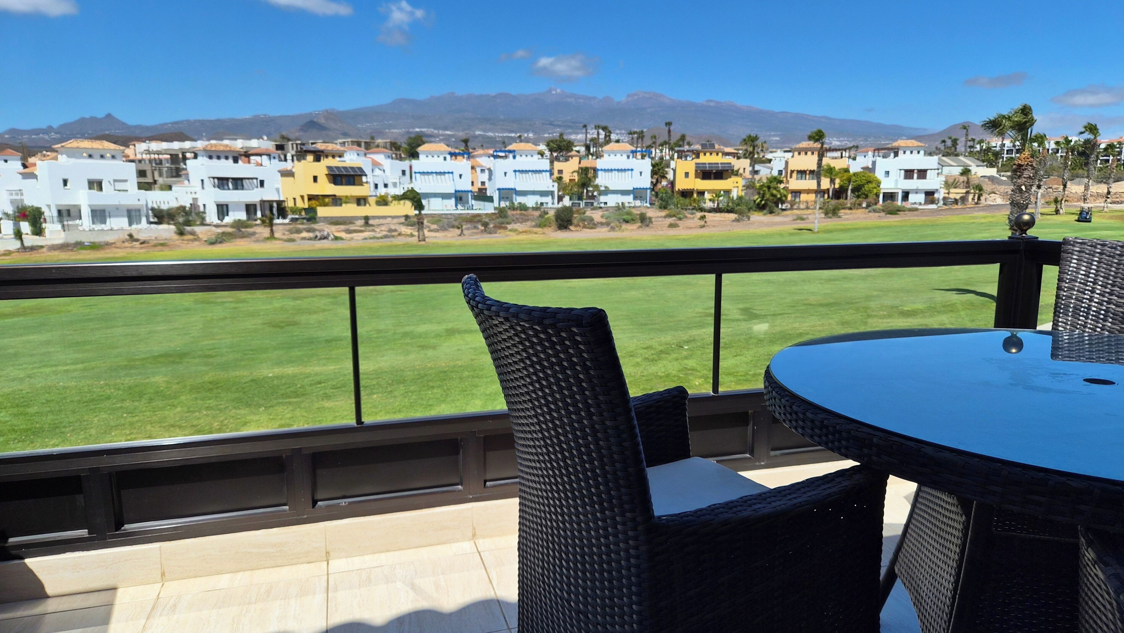 A balcony with wicker chairs and a table, overlooking a green lawn and a row of colorful buildings with mountains in the background.