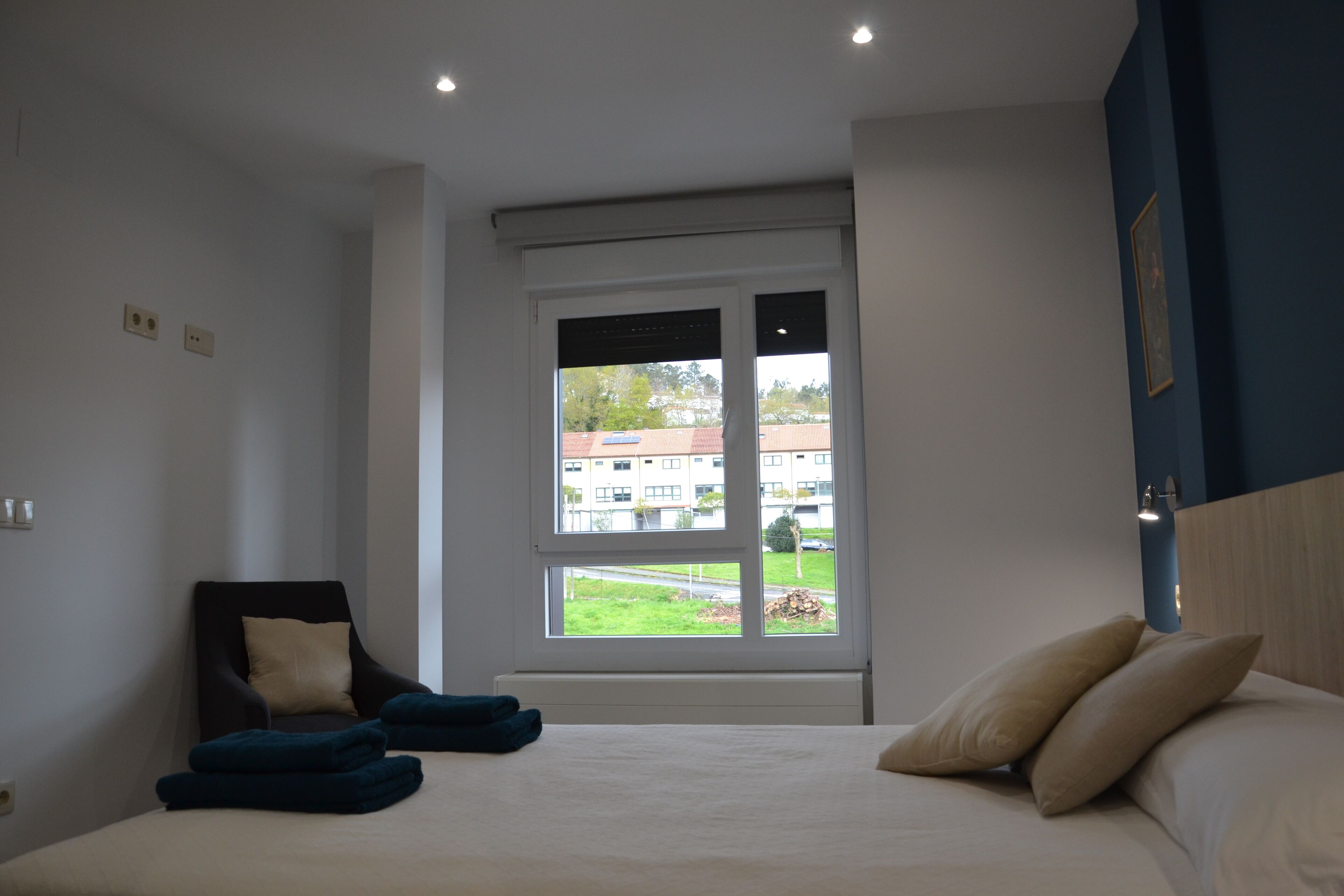 Un dormitorio con cama, una silla y ventana con vistas a edificios y follaje.
