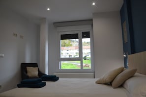 Ein Schlafzimmer mit Bett, Sessel und einem Fenster mit Blick auf Gebäude und Grünflächen.