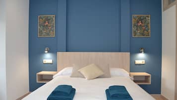 Una cama bien hecha con dos toallas dobladas, un cabecero de madera, dos mesitas de noche fijadas a la pared y dos luces fijadas a la pared.