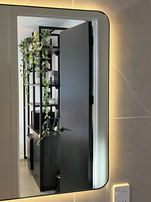 A mirror reflecting a room with a dark-colored cabinet, a plant, and a shelf with various items. - Modern and Peaceful Studio Room + Ensuite Bathroom under Mountains! (Jack's Point)