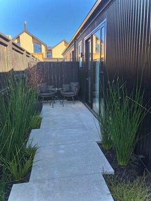 A modern outdoor patio with a concrete pathway, two grey cushioned chairs, and tall green plants. - Modern and Peaceful Studio Room + Ensuite Bathroom under Mountains! (Jack's Point)