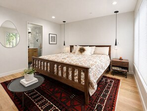 Ein Schlafzimmer mit einem hölzernen Bett, einem runden Tisch mit einer Pflanze, einem schwarz-roten Teppich, einem Nachttisch und einem Fenster mit Jalousien.