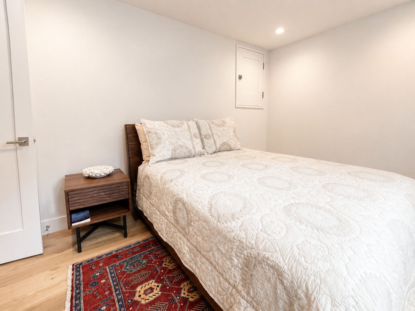 Une chambre à coucher avec un lit, une table de chevet, un tapis rouge à motifs et une porte blanche.