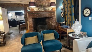 Een woonkamer met een metselwerk openhaard, blauwe fauteuils, een witte bank en een eethoek op de achtergrond.